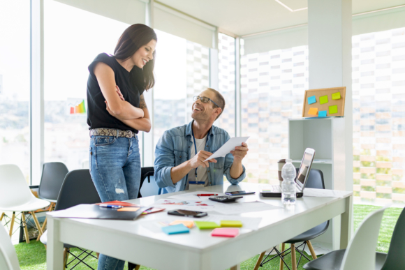 Man showing off brainstorming handiwork to female co-worker standing over his shoulder with arms folded together.